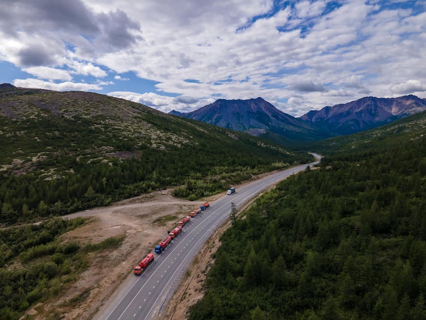 Aerial shot of a mountain highway with trucks, surrounded by lush greenery and distant mountains under a cloudy sky.